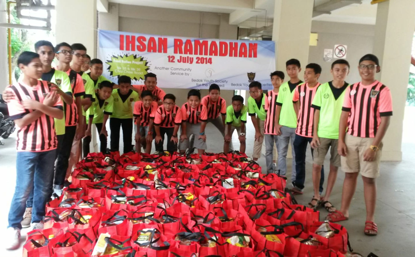 A group of boys in striped and neon green jerseys pose around a large number of red bags filled with supplies under a banner reading “IHSAN RAMADHAN – 12 July 2014,” showing a community service effort.