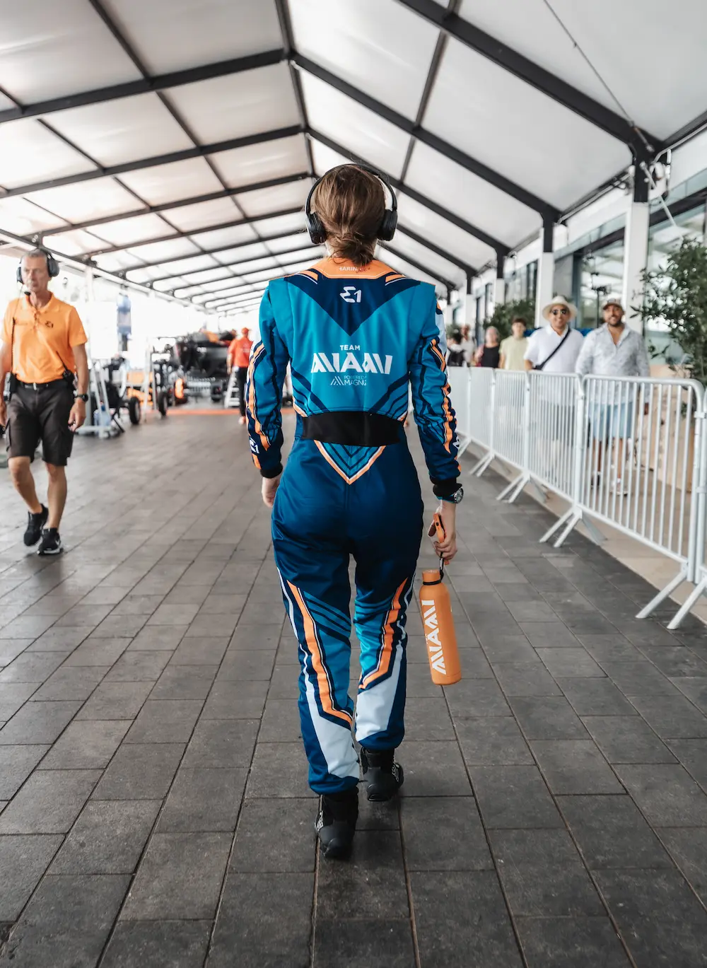E1 Team Miami racer walks confidently through the paddock in full branded Marina Racewear suit, holding a matching orange water bottle during race day preparations.