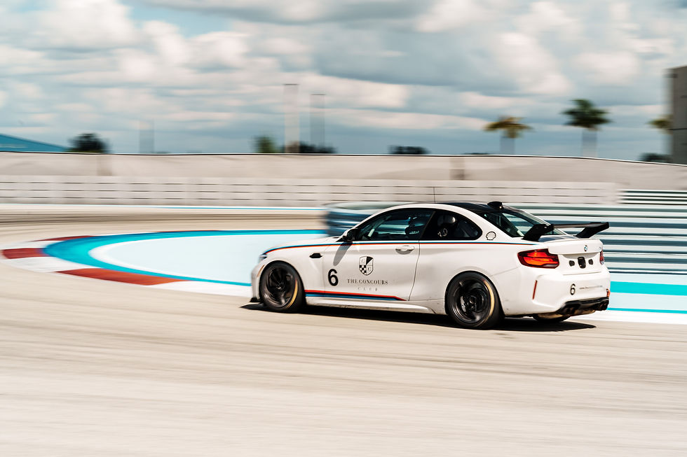 White BMW race car with number 6 and The Concours Club logo speeds around a track under cloudy skies, with motion blur.