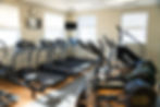 Bright gym room with treadmills, ellipticals, and other fitness equipment. Windows and a clock on beige walls. Calm, clean atmosphere.