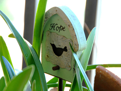 A charming sign in the style of a birdhouse is nestled amidst the green leaves of a plant, the word "Hope" drawn on it in cursive.