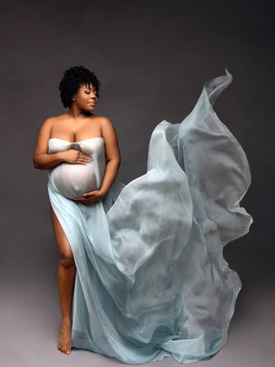 Pregnant dark-skinned woman draped in sheer light blue flowing fabric cradling bump in side profile against deep blue studio backdrop — Long Island maternity photographer — Imagine That Photography, Dix Hills NY