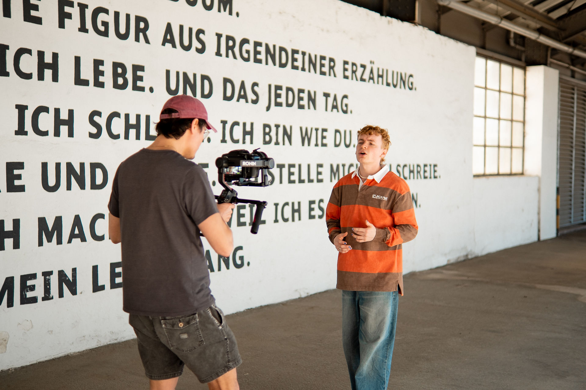 Ein Künstler tritt in die Kamera, die vor einer weißen Wand mit schwarzem Text gefilmt wird.