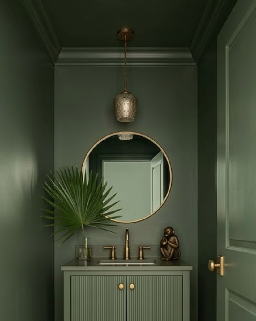 Elegant green bathroom with a round mirror, palm leaf, bronze fixtures, and a small monkey statue on a cabinet. Mood: serene.