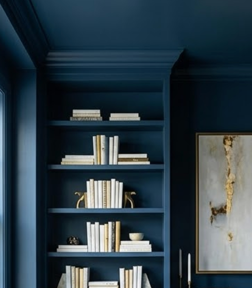 Navy room with a built-in bookshelf filled with white and gold books, decorative objects. Abstract painting on wall, calm atmosphere.