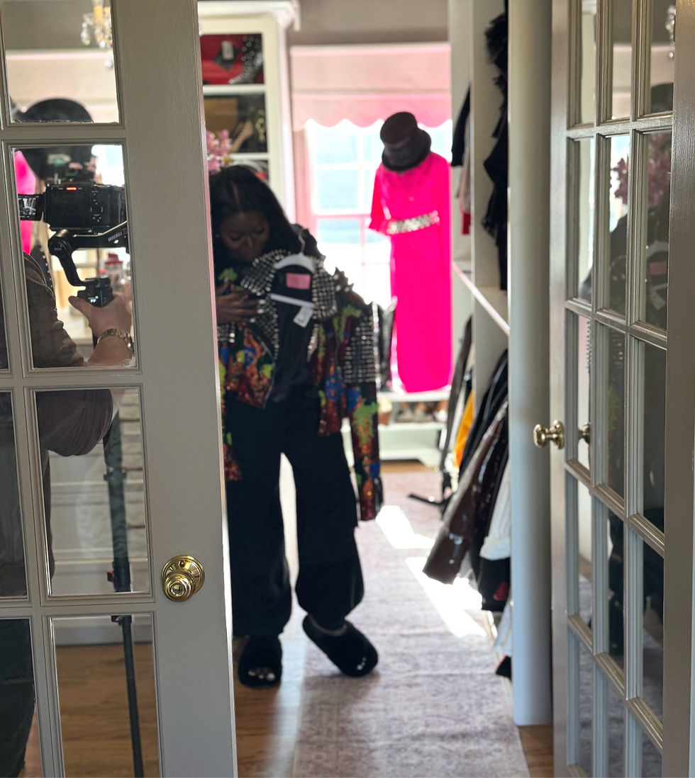 A person stands in a room holding colorful clothes, reflected in a glass door. A camera is visible. A pink dress hangs in the background.