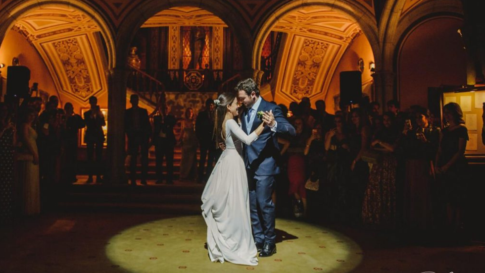 A couple dances in a warmly lit, ornate hall surrounded by guests. The woman wears a white dress, the man a suit. Romantic and elegant mood.