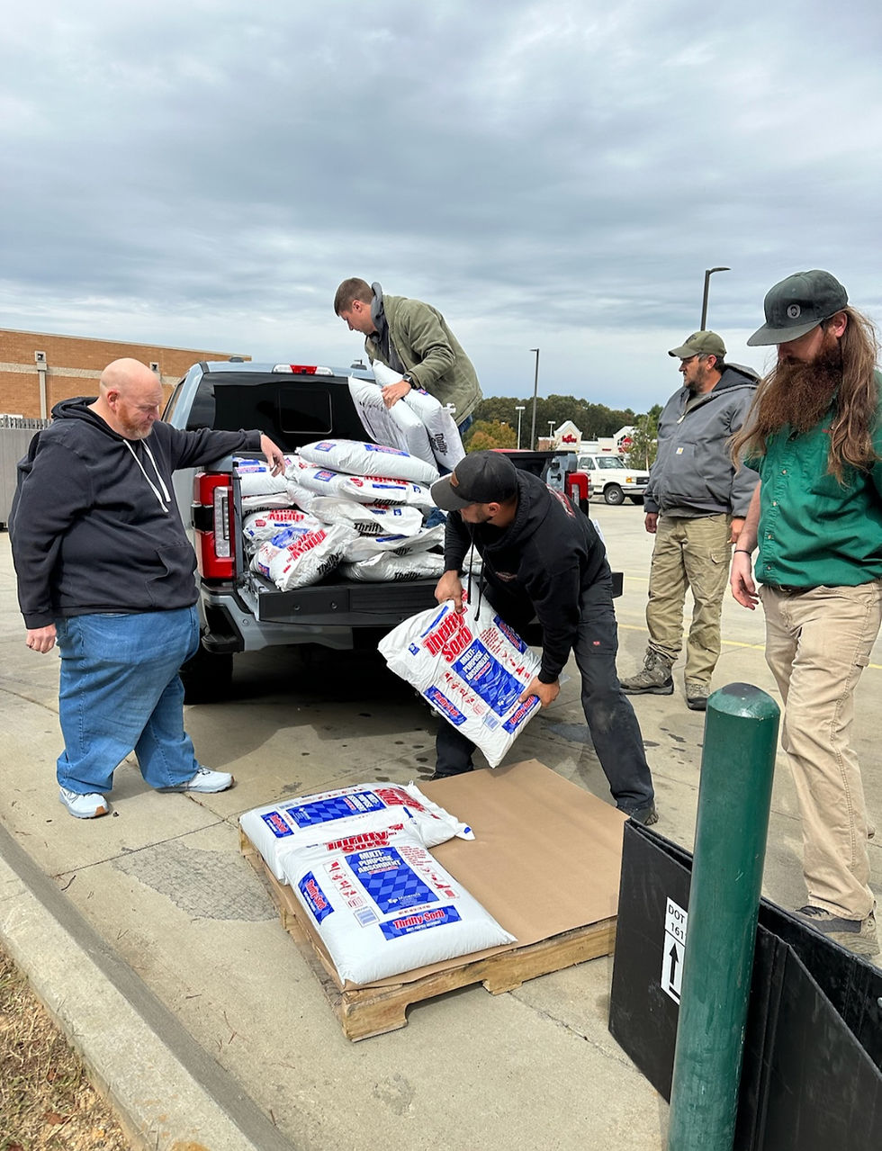 Five men unload white bags of pellets from a truck onto a wooden pallet in a parking lot. Overcast sky background.