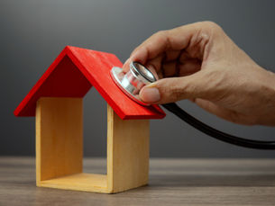 A hand holds a stethoscope to the roof of a small wooden house, representing the concept of home inspection or property assessment