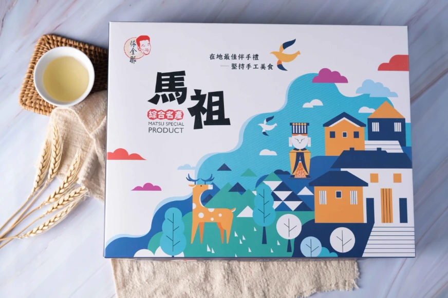 包裝盒上印有彩色圖案和文字“馬祖綜合名產”，背景為卡通風格房屋、鹿和飛鳥，旁邊是小麥和茶杯。