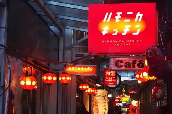 狹窄街道上方掛滿紅燈籠，充滿溫暖氛圍，多個紅色招牌寫著​​HARMONICA KITCHEN AND BAR和其他日文，背景是夜晚的城鎮景色。