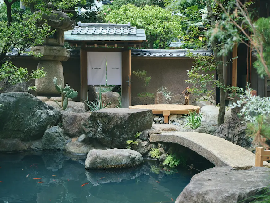 寧靜的日式庭院，池塘中有紅色錦鯉，石橋上方是綠色植物和苔蘚，大石旁有一塊寫有