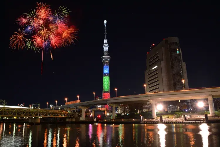 夜晚的隅田川花火大會，東京晴空塔被彩燈照亮，背景煙火絢麗綻放。城市景觀與水面倒影增添節慶氣氛，天空漆黑無雲。