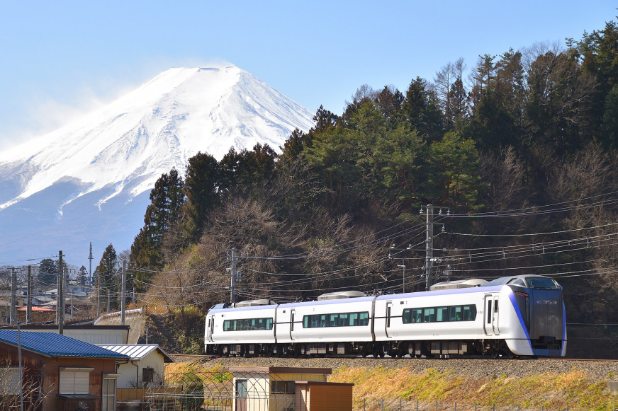 火車行駛在鐵軌上，背景是藍天白雪覆蓋的富士山，周圍綠色森林環繞，顯示出寧靜的鄉村風景。