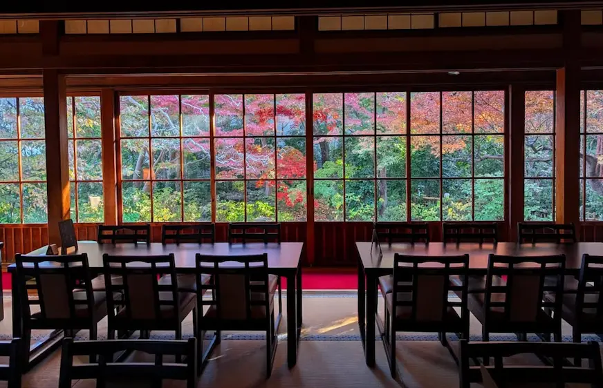日式房間內，黑色桌椅整齊排列。窗外秋葉紅黃交織，景色優美。室內光線柔和，營造寧靜氛圍。