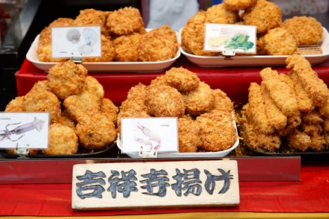三個托盤裝滿日式油炸小吃，放在紅色背景上。每個托盤上有插牌，底部有「吉祥寺揚げ」木牌。食物呈金黃色。