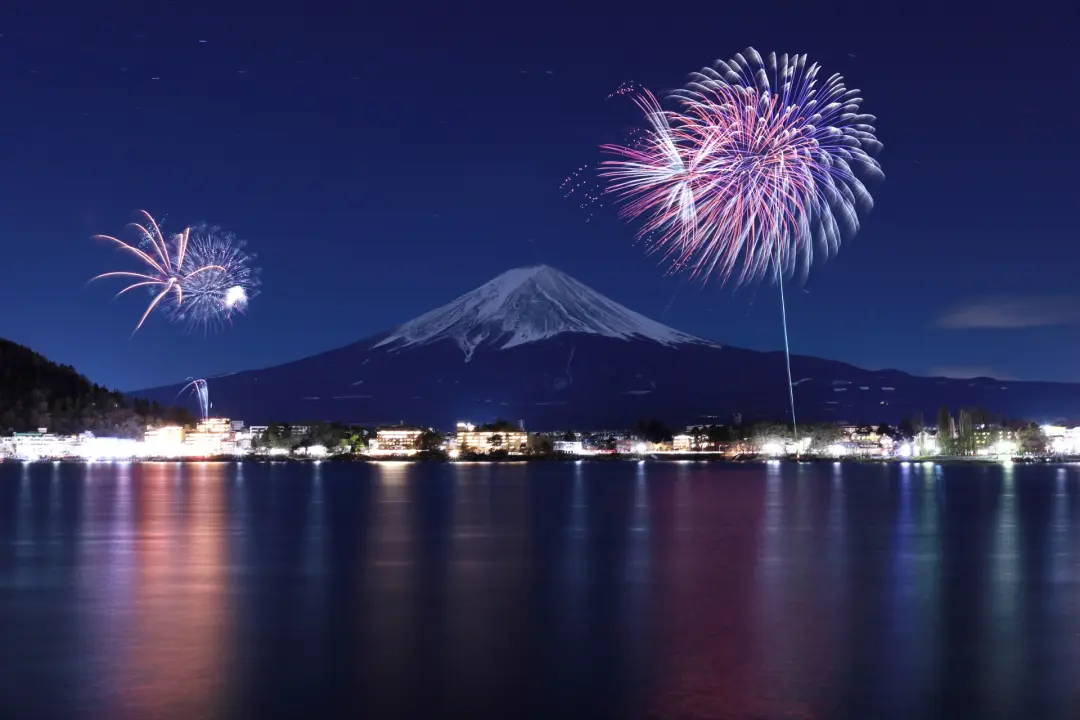 河口湖冬花火，夜空中煙火綻放，遠處富士山靜謐矗立，湖面倒映城市燈光，營造出寧靜浪漫的節慶氛圍。