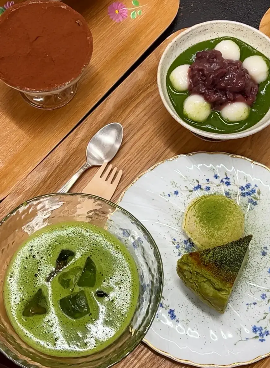 木桌上擺放著抹茶甜點。包括紅豆白玉團、抹茶慕斯蛋糕和抹茶飲料，旁邊有湯匙和叉子，呈現日式風格。