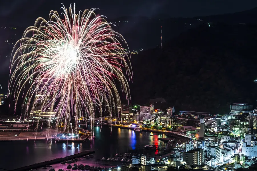 熱海海上花火大會，夜晚城市的煙火秀：璀璨的煙火在夜空中綻放，城市燈火輝煌，倒映在水面上，色彩鮮豔