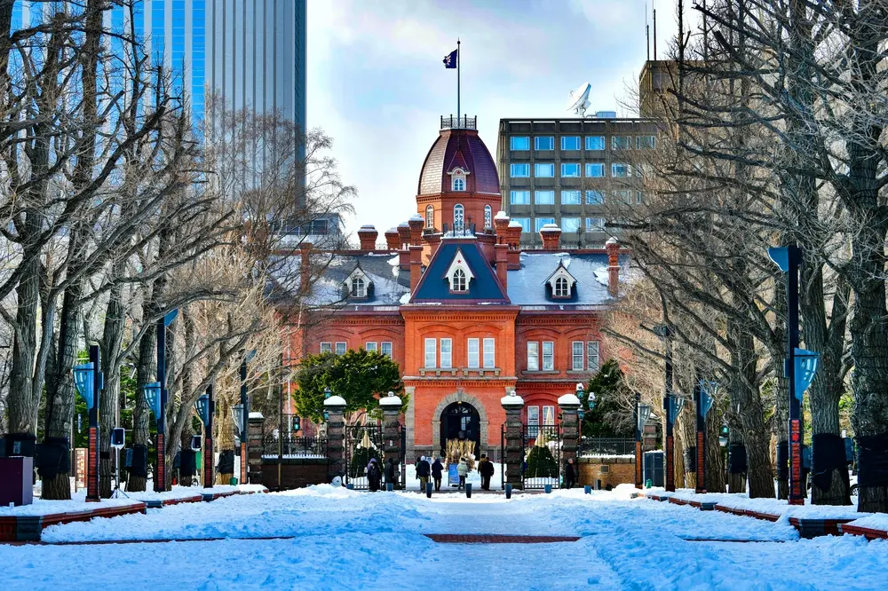 雪地中的紅磚歷史建築，中央塔樓和尖頂屋頂，前方有人步行，四周落葉樹木與藍色路燈，背景是現代高樓，冬日寧靜景象