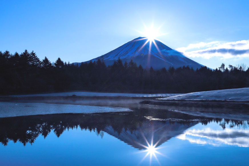 藍天下富士山頂日光閃爍，映射在平靜的湖面上。湖邊樹影婆娑，畫面寧靜唯美。