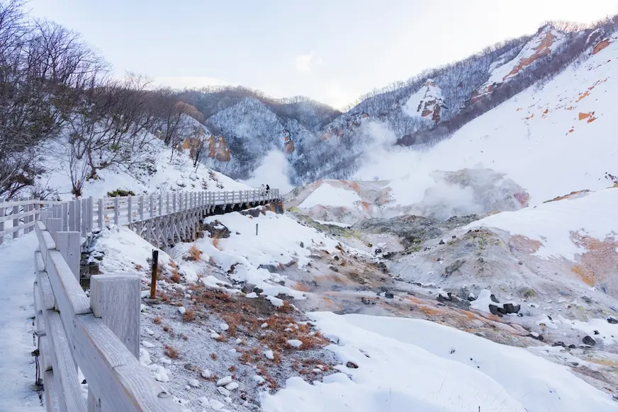 雪覆山谷中的地熱景區，木棧道沿坡延伸，蒸汽從岩地噴出，遠處群山靜謐寒冷。