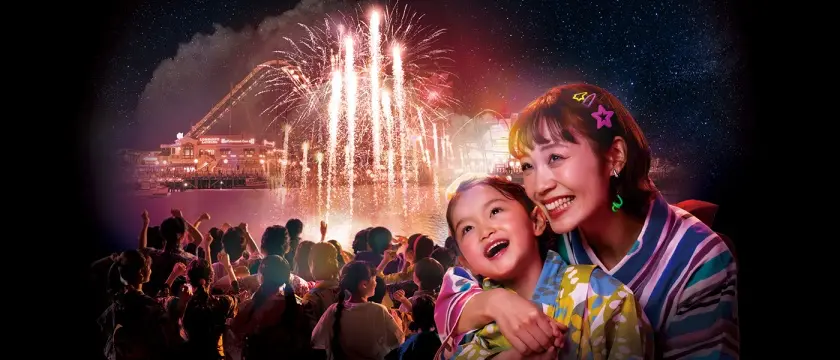 夜晚煙花秀前，穿彩色和服的女子擁抱笑著的小女孩，身後人群仰望湖面煙花和燈光，氛圍歡樂熱鬧。