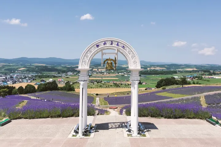 白色拱門前俯瞰紫色薰衣草花田與綠色鄉野，遠處村鎮和群山在晴朗藍天下，景色開闊寧靜。