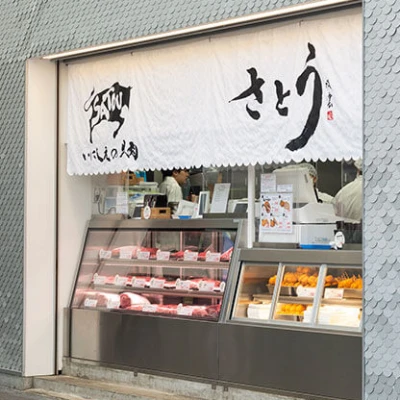 商店外觀，展出肉類和油炸食品，窗簾上有「さとう」字樣，內部可見員工在工作，氣氛繁忙。