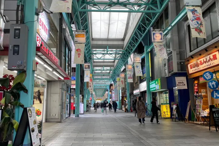 在商店街的拱廊內，人們在走動，周圍是色彩鮮豔的店鋪招牌和廣告，天花板採用白色和綠色框架結構，氛圍繁忙。