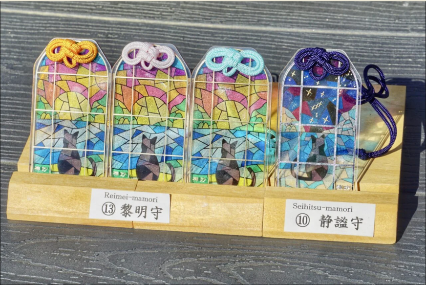 木架上陳列四枚彩色和風護身符，分別系黃、白、淺藍、深紫繩結，圖案像彩窗與黑貓，標籤寫黎明守、靜謐守，氣氛安靜雅致