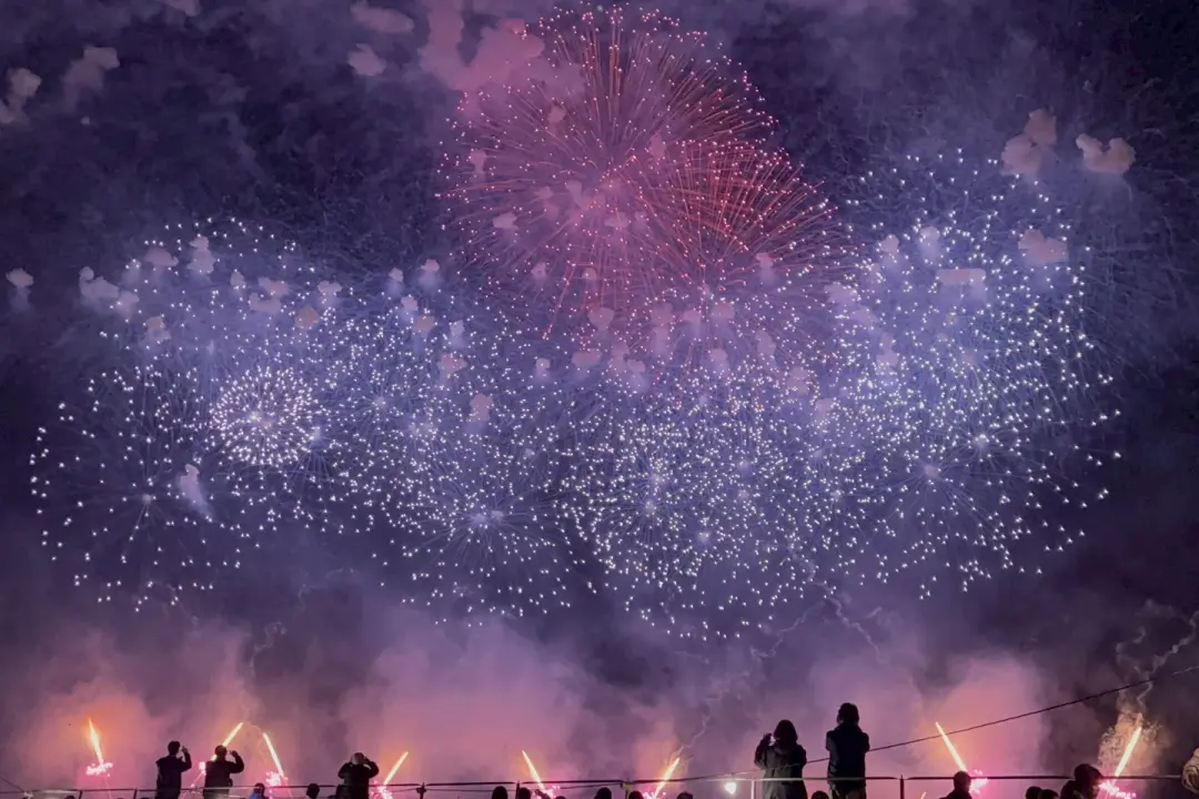 土浦全國花火競技大會煙火在夜空中綻放，呈現紫色和粉紅色。下方幾個人拍照觀賞，氣氛熱鬧歡慶。