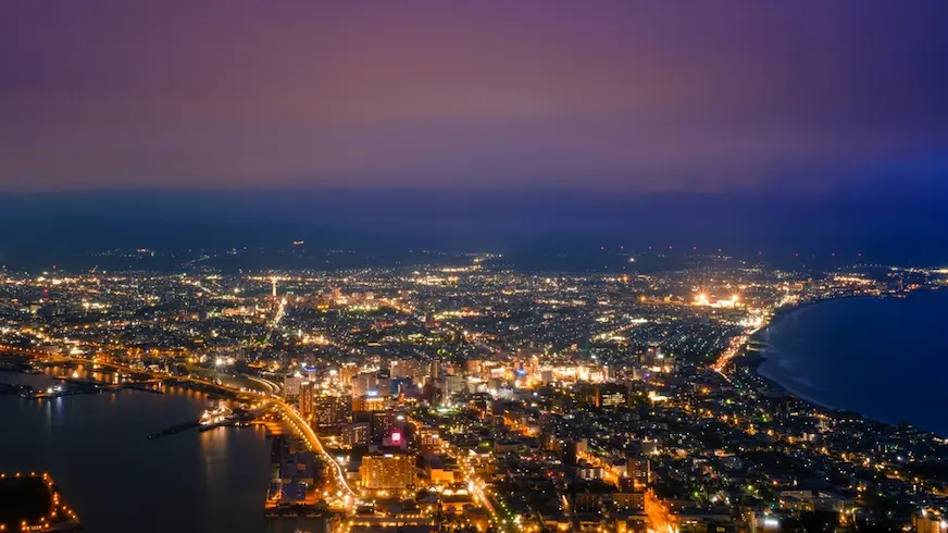 函館夜景航拍海濱城市，港灣與海岸線清晰可見，密集燈火點亮街區與道路，遠處天際泛紫，氛圍靜謐璀璨