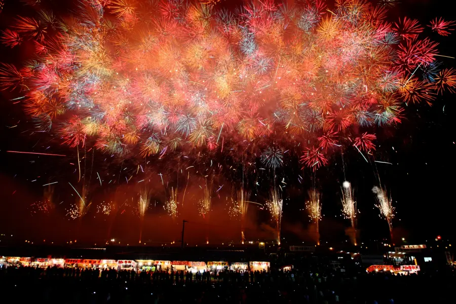 大曲之花火，夜空中絢麗煙火綻放，紅色和金色光芒交織。下方有觀眾和亮燈的攤位，營造出歡樂慶典的氣氛。