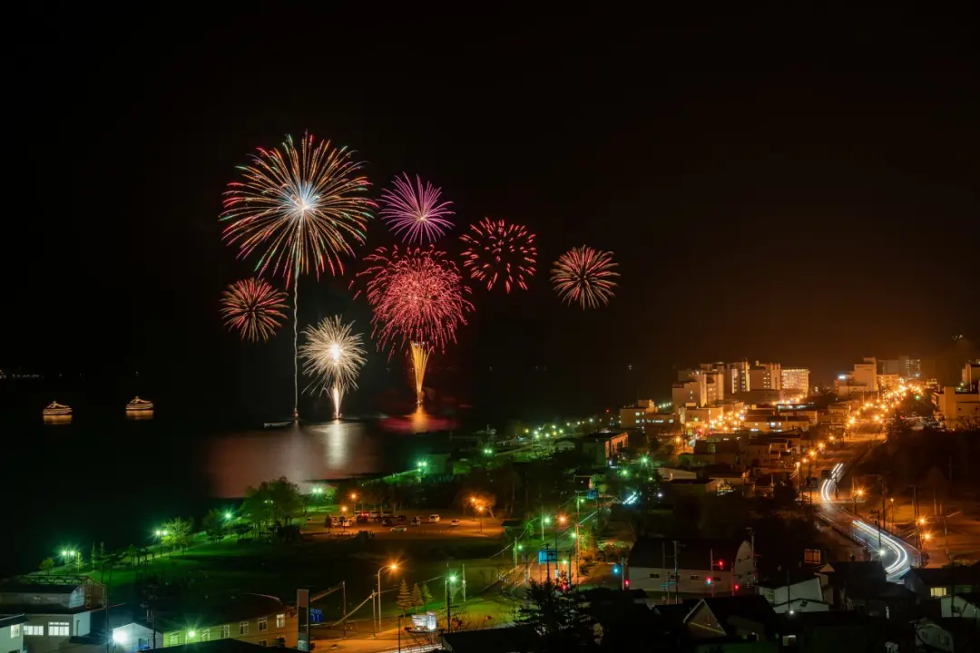 洞爺湖花火大會期間，夜空中綻放五彩繽紛的煙火，映照著旁邊城市的燈火。煙火倒映在水中，畫面充滿節慶氛圍。