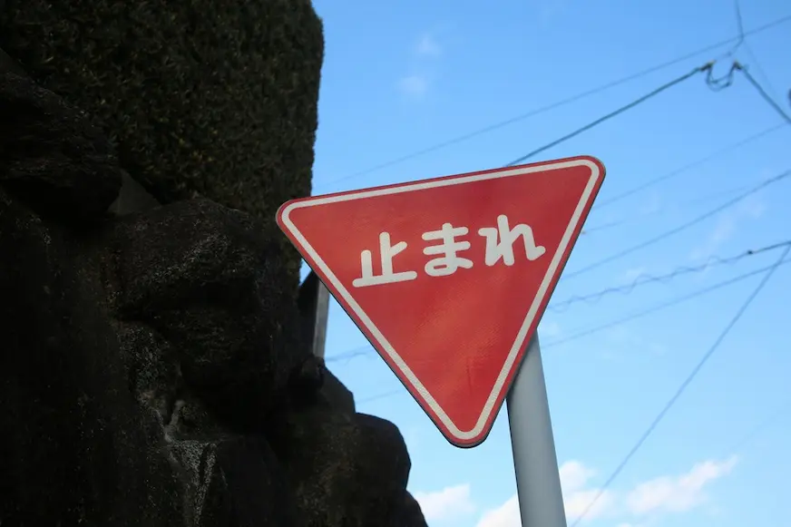 紅色倒三角停車讓行標誌，上面寫著“止まれ”，豎立在藍天和電線前，左側有深色樹影／岩壁。