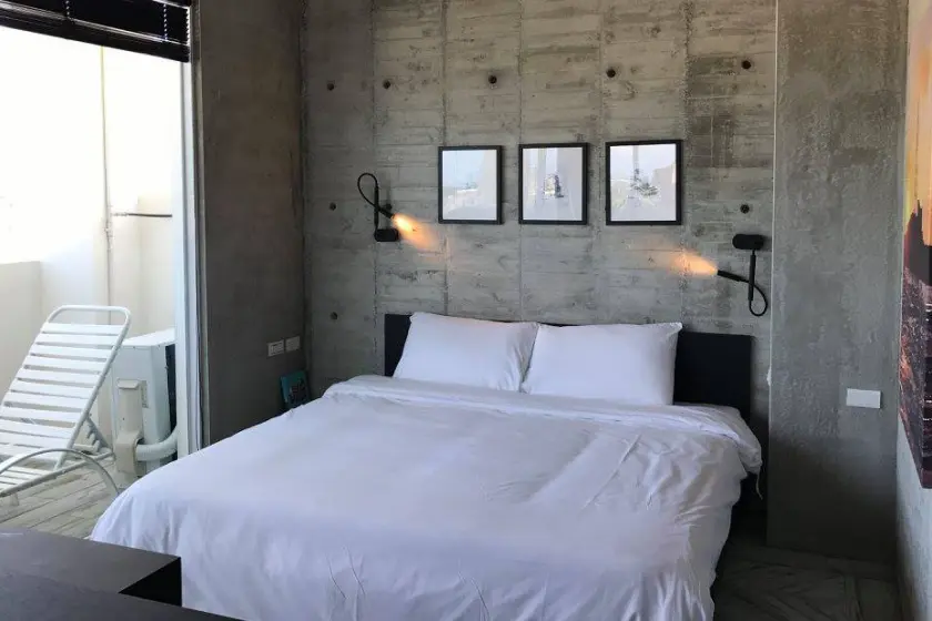 彩色珊瑚礁宿，房內白色床鋪靠在混凝土牆上，牆上有三幅裝飾畫，床兩側有壁燈，窗外有躺椅，簡約風格。