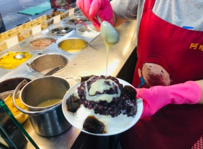 穿紅色圍裙的店員正給一碗剉冰澆煉乳，剉冰上有紅豆和仙草塊，背景有調料盤，