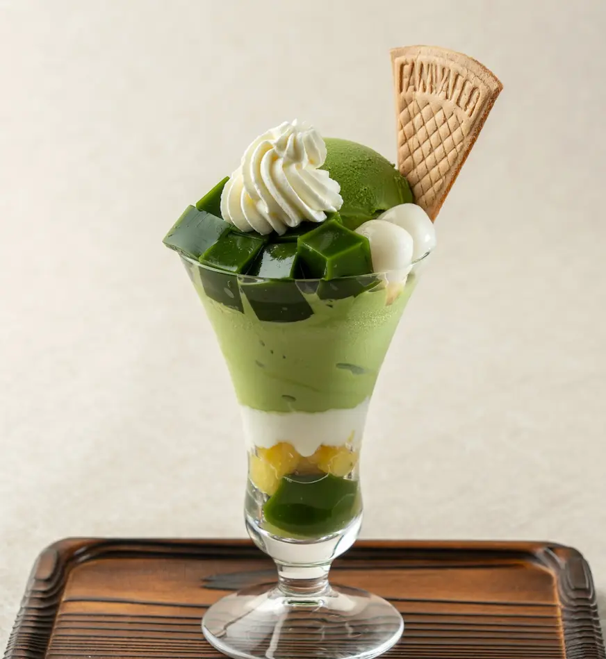 透明玻璃杯內盛裝抹茶冰淇淋、奶油、綠茶凍、白玉團子和曲奇餅乾，置於木托盤上，整體色彩鮮豔誘人。