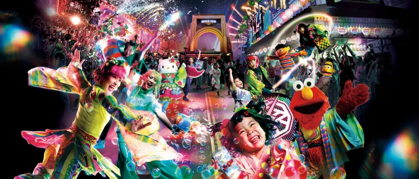 夜晚的遊行現場，演員和Hello Kitty、Elmo等角色在彩色燈光、泡泡和紙屑中歡快舞動，背景有“UNIVERSAL”拱門，氣氛熱鬧狂歡。
