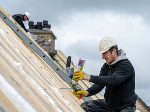 Roofer in hard hat applies sealant to a sloped roof while another worker stands nearby under a cloudy sky.