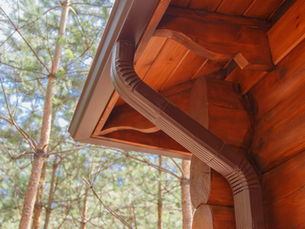 Close-up of a wooden cabin eave with brown gutter and downspout, framed by sunlit pine trees in the background.