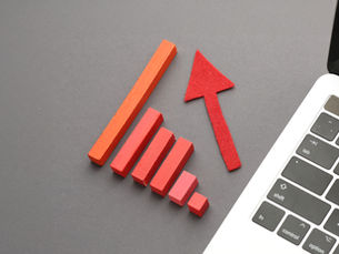 Red bar chart with upward arrow beside a laptop keyboard on a gray desk, suggesting growth or rising trends.