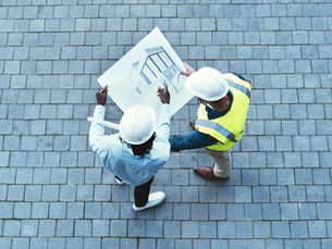 Two construction workers in hard hats review a building blueprint on a gray stone-paved site, viewed from above.