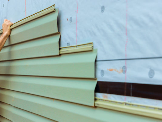 Hand installing beige vinyl siding on a house wall over white wrap, with pink guide lines and circular markers.