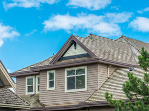 Suburban house roof and windows beneath a bright blue sky with scattered clouds, framed by a pine tree