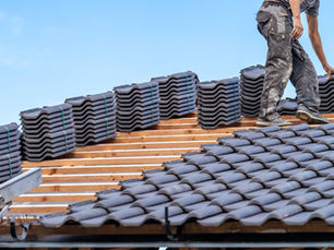Worker installing black roof tiles, standing on wooden beams under a clear blue sky. Neatly stacked tiles in foreground.