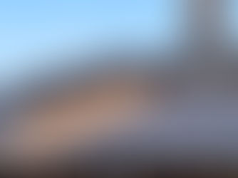 Worker installing black roof tiles, standing on wooden beams under a clear blue sky. Neatly stacked tiles in foreground.