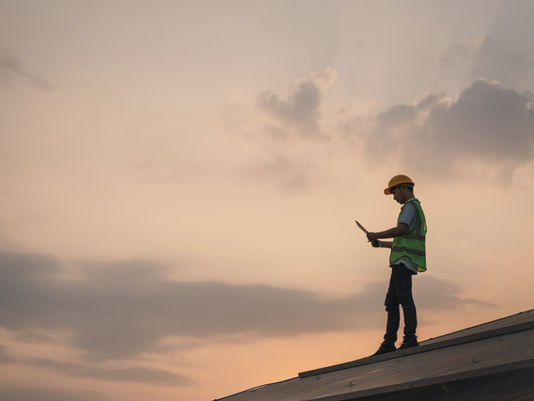 A construction worker in a safety vest and hard hat stands on a rooftop using a tablet at sunset, with cloudy skies in the background.