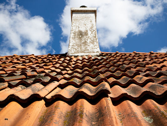 Old terracotta tile roof with a white chimney under a blue sky with scattered clouds.